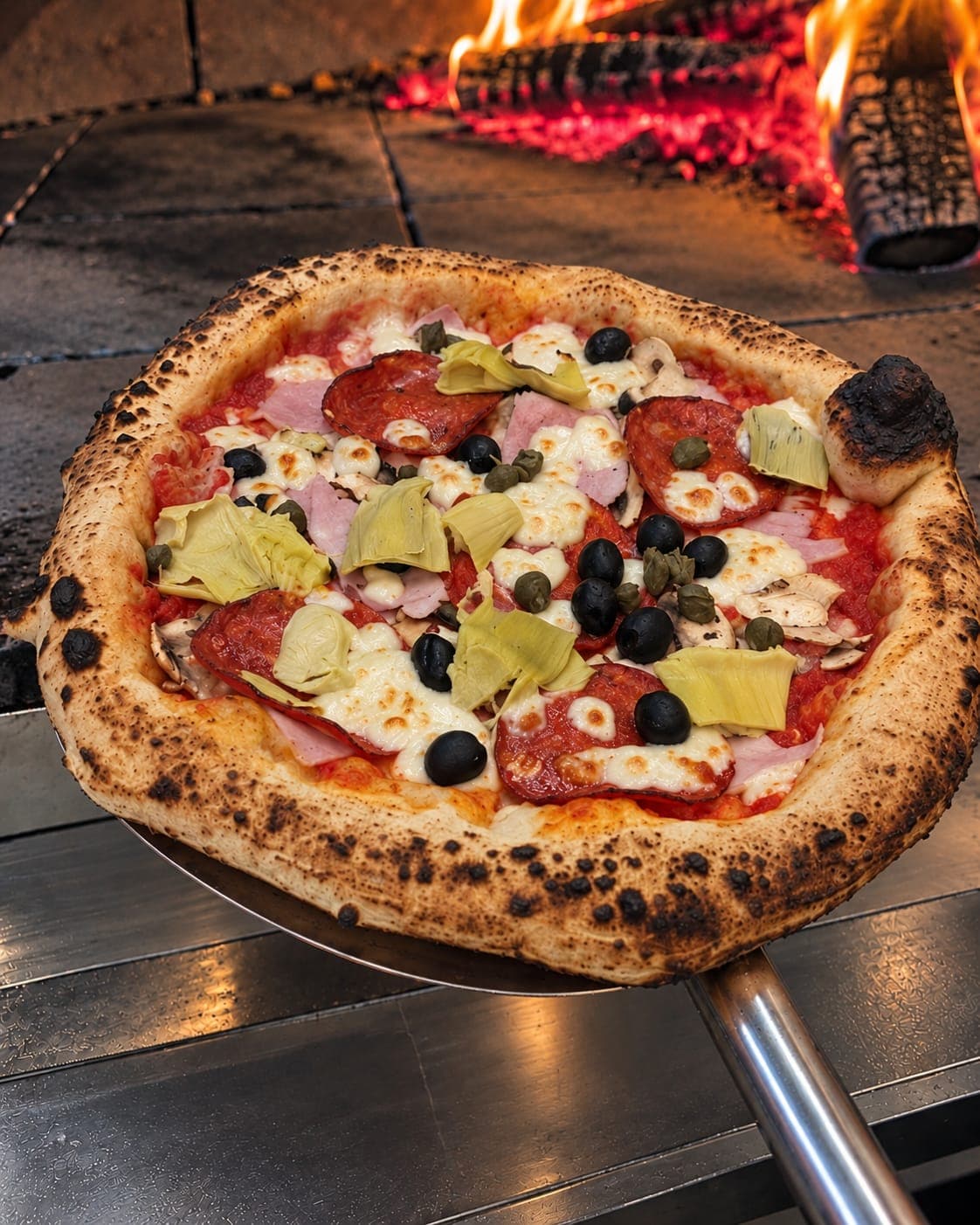 La pizza 4 saisons sur la pelle, devant les flammes du four à bois.