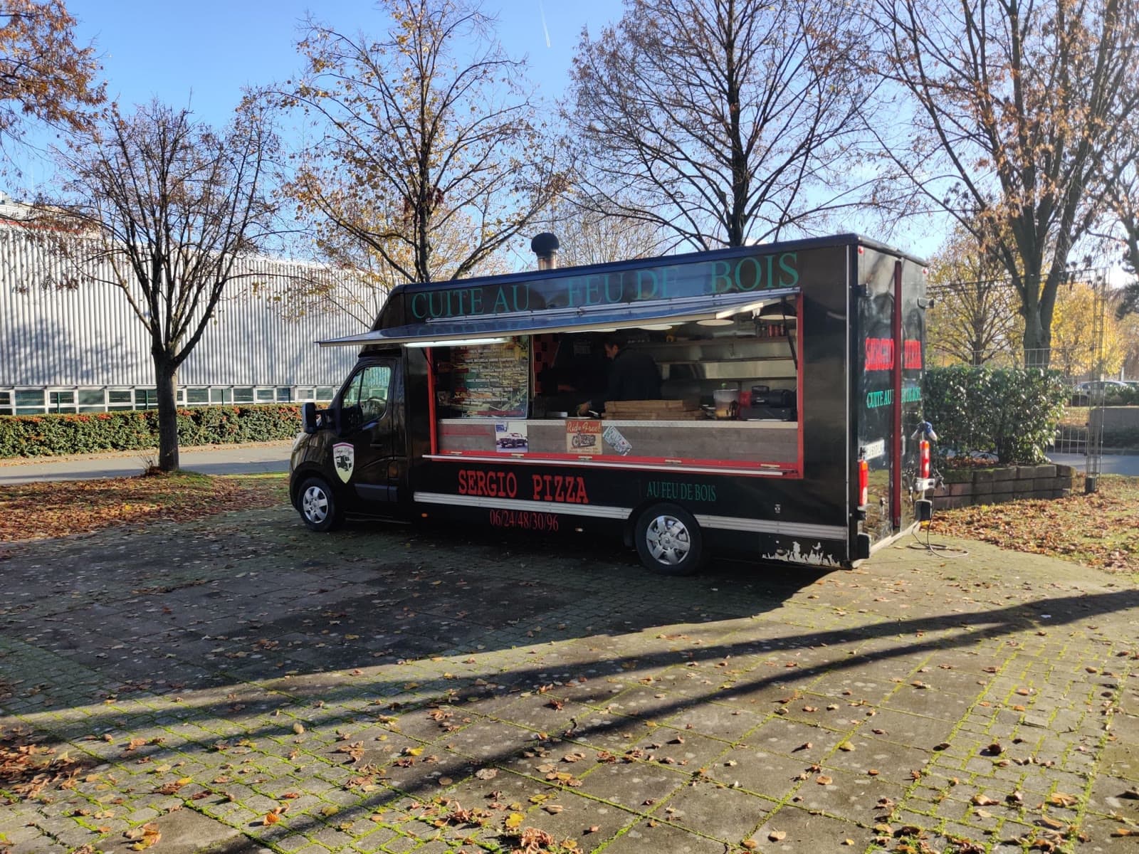 Le food truck Sergio Pizza ouvert sur une place pavée, sous les arbres d'automne.