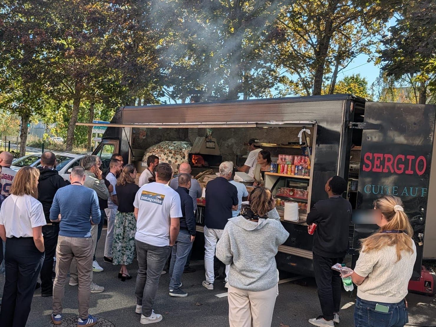 Une trentaine de personnes font la queue devant le camion ouvert, le four allumé, sous les arbres.