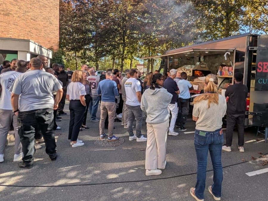 Vu de dos, un groupe attend devant le camion Sergio Pizza, la fumée du four monte dans les arbres.