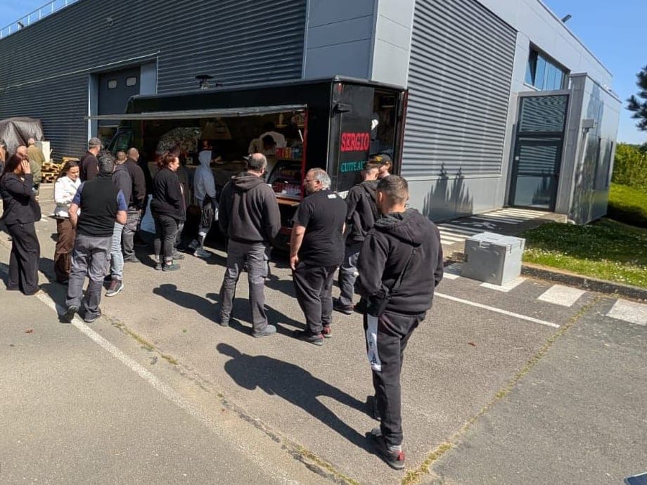 Le camion installé devant un bâtiment gris, des invités servis au comptoir sur le parking.