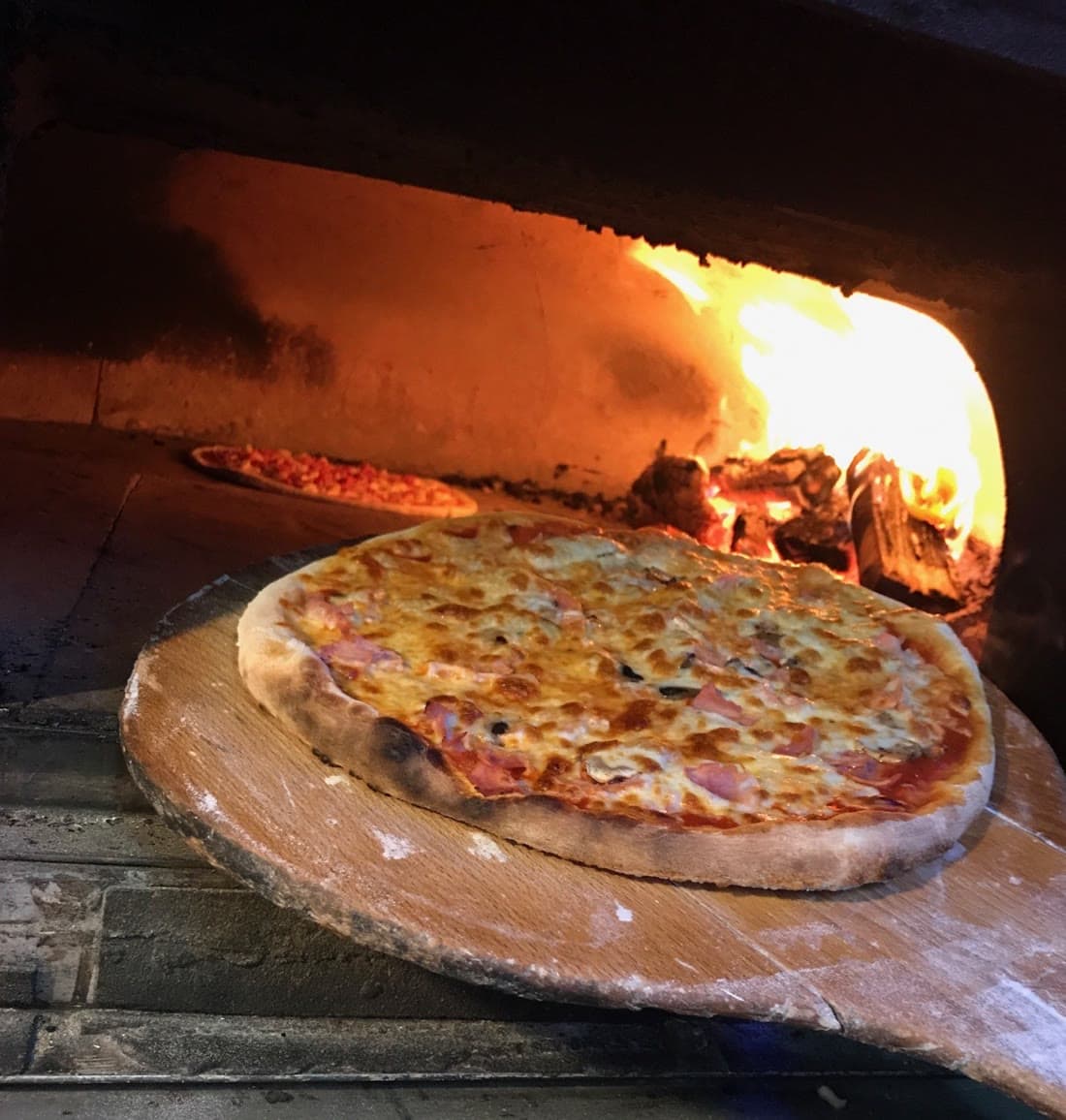 Une pizza au jambon sur la pelle en bois, à la sortie du four, les flammes derrière.